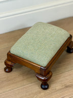 A small wooden footstool with a light sage green herringbone upholstered cushion, shown on a light wood floor against a white wall.
