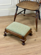 A small wooden footstool with a light sage green upholstered cushion, shown on a light wood floor next to a larger wooden chair.