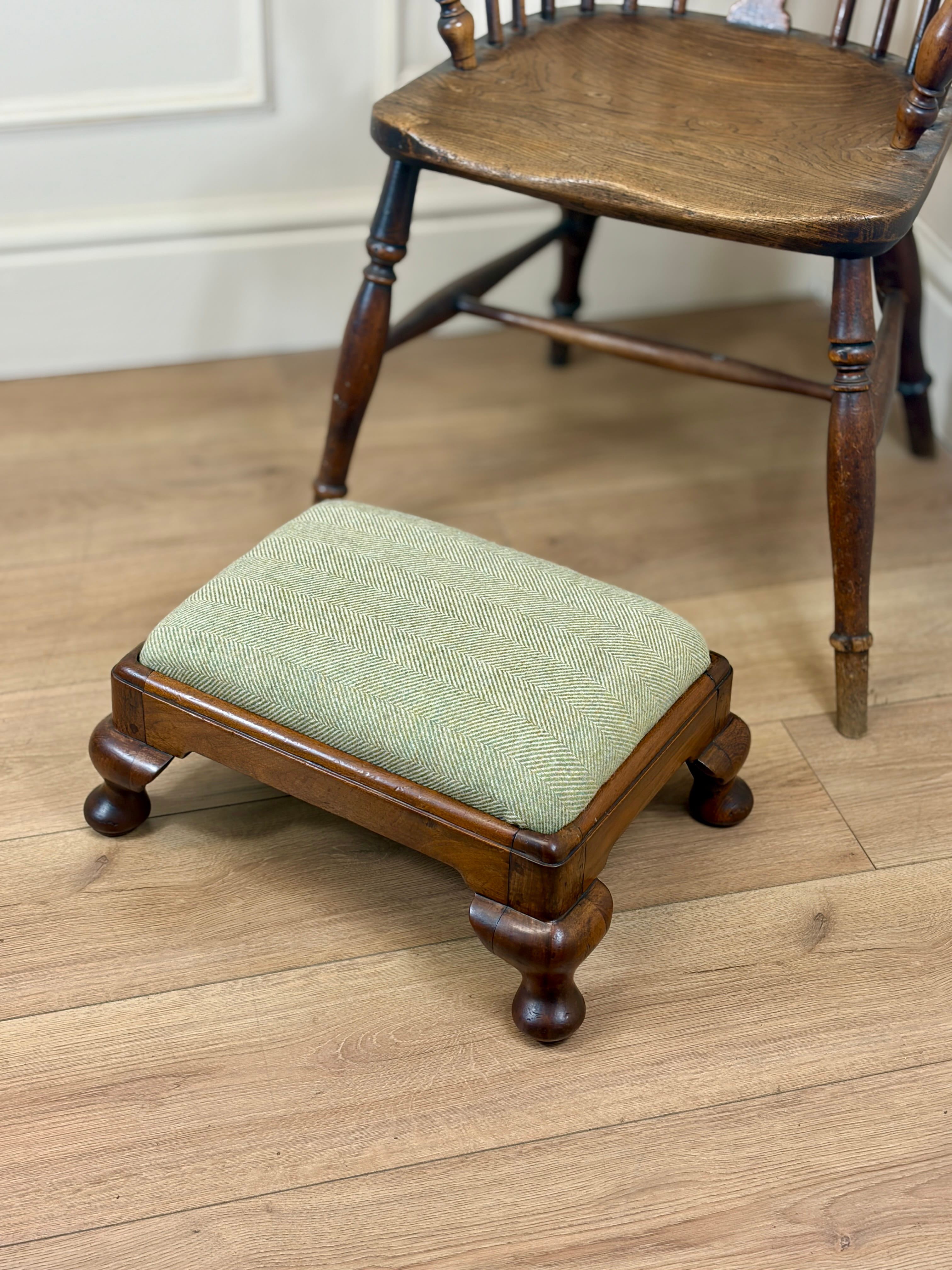 A small wooden footstool with a light sage green upholstered cushion, shown on a light wood floor next to a larger wooden chair.
