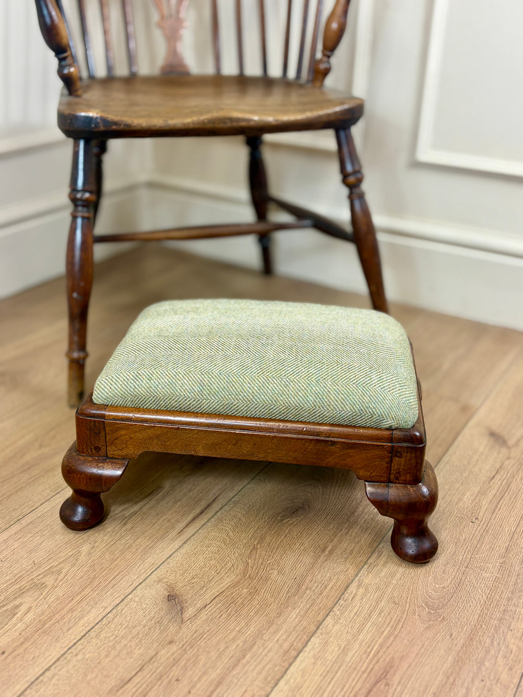 A small wooden footstool with a light sage green herringbone upholstered cushion, positioned in front of a wooden spindleback chair on a light wood floor.