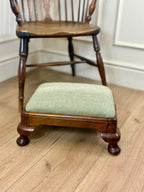 A small wooden footstool with a light sage green herringbone upholstered cushion, positioned in front of a wooden spindleback chair on a light wood floor.