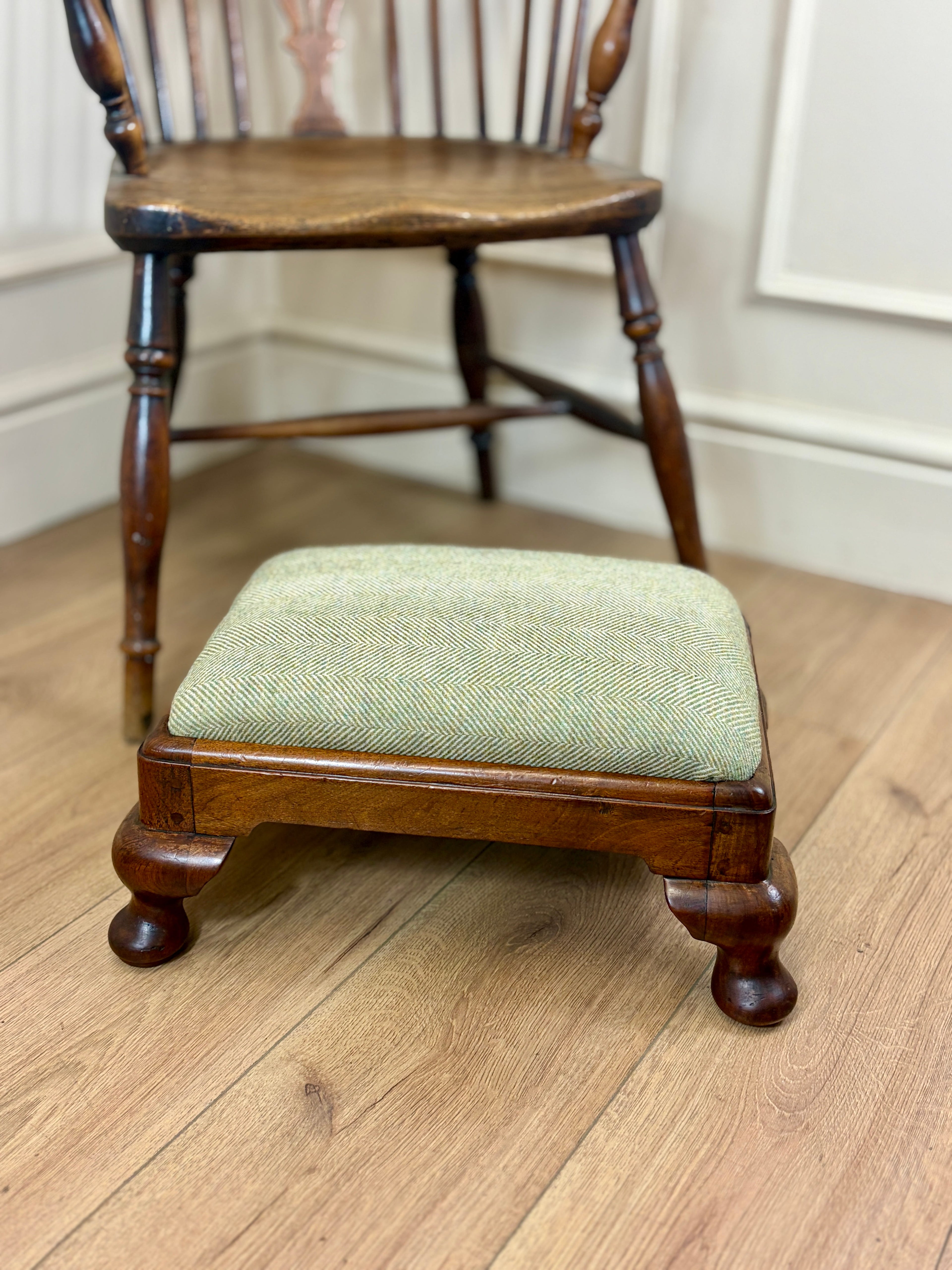 A small wooden footstool with a light sage green herringbone upholstered cushion, positioned in front of a wooden spindleback chair on a light wood floor.