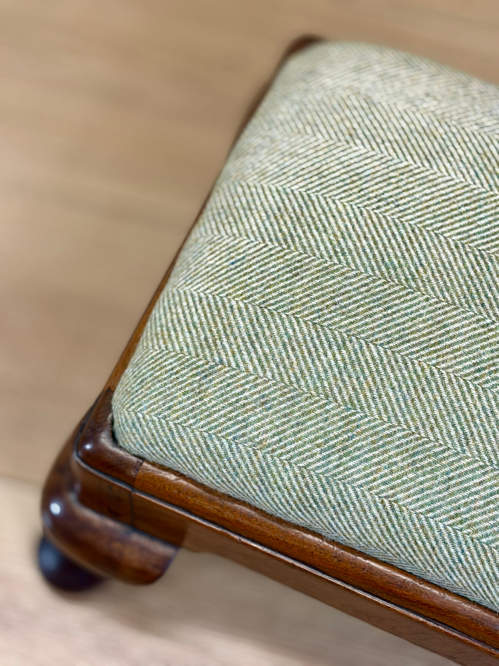 Close-up of a wooden stool or bench with a light sage green and beige herringbone upholstered seat cushion, set against a light wood floor background.