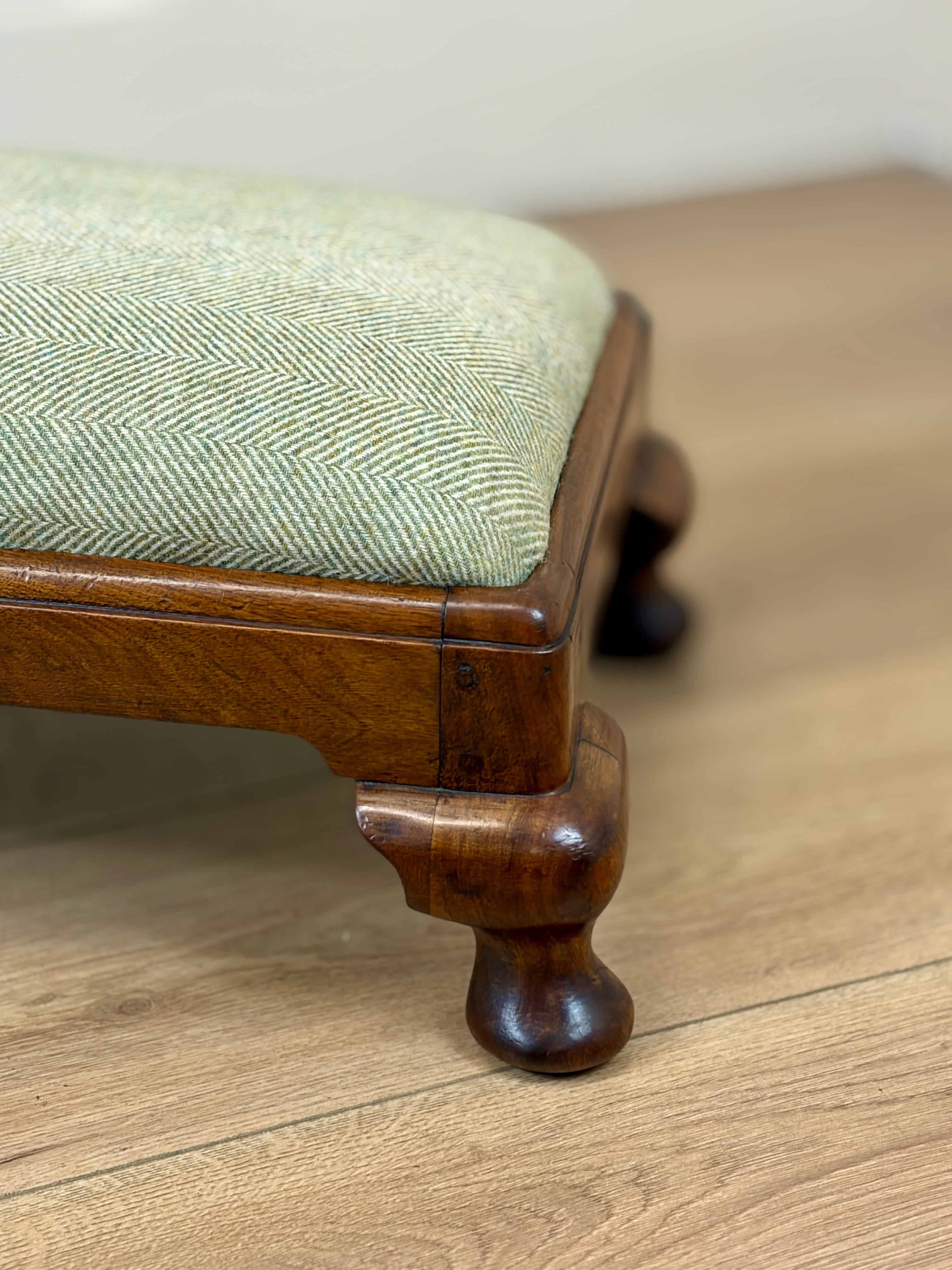 Close-up of a wooden stool or bench with a light sage green herringbone upholstered seat cushion and turned wood legs, shown on a light wood floor.