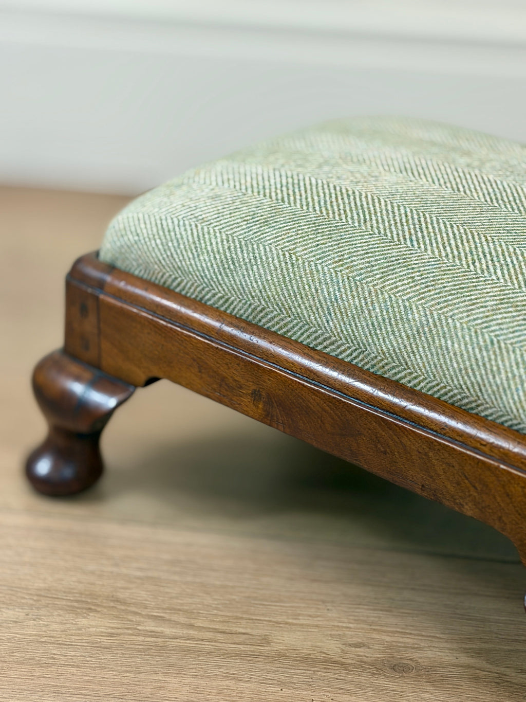 A close-up of a wooden stool or ottoman with a light sage green herringbone upholstered seat, resting on a light wood floor.
