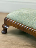 A close-up of a wooden stool or ottoman with a light sage green herringbone upholstered seat, resting on a light wood floor.