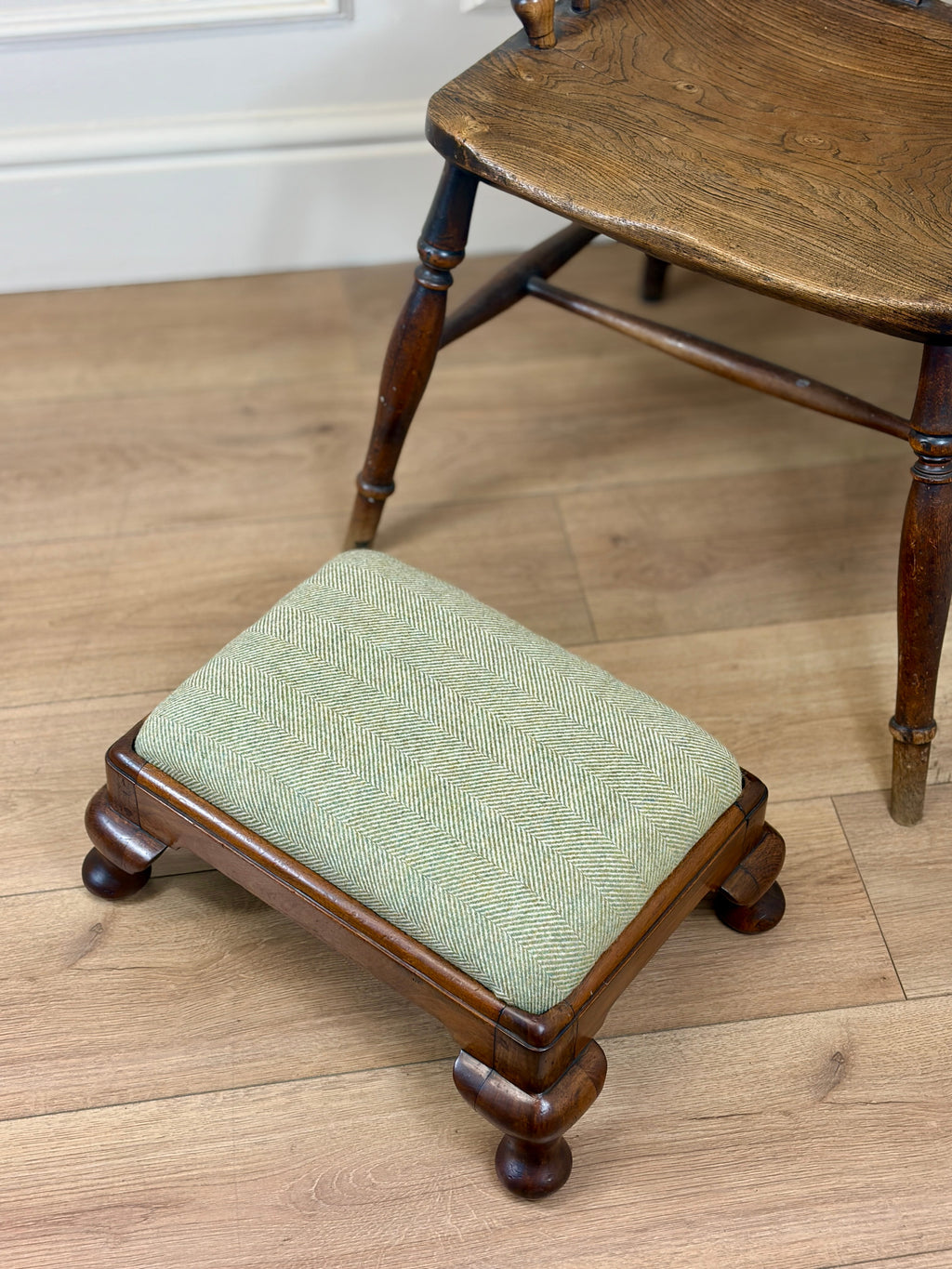 A small wooden footstool with a light sage green herringbone upholstered cushion, shown on a light wood floor next to a larger wooden chair.