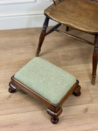 A small wooden footstool with a light sage green herringbone upholstered cushion, shown on a light wood floor next to a larger wooden chair.