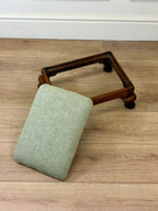 A small upholstered cushion in a light sage green fabric rests against a dark wood rectangular frame on a light oak floor.
