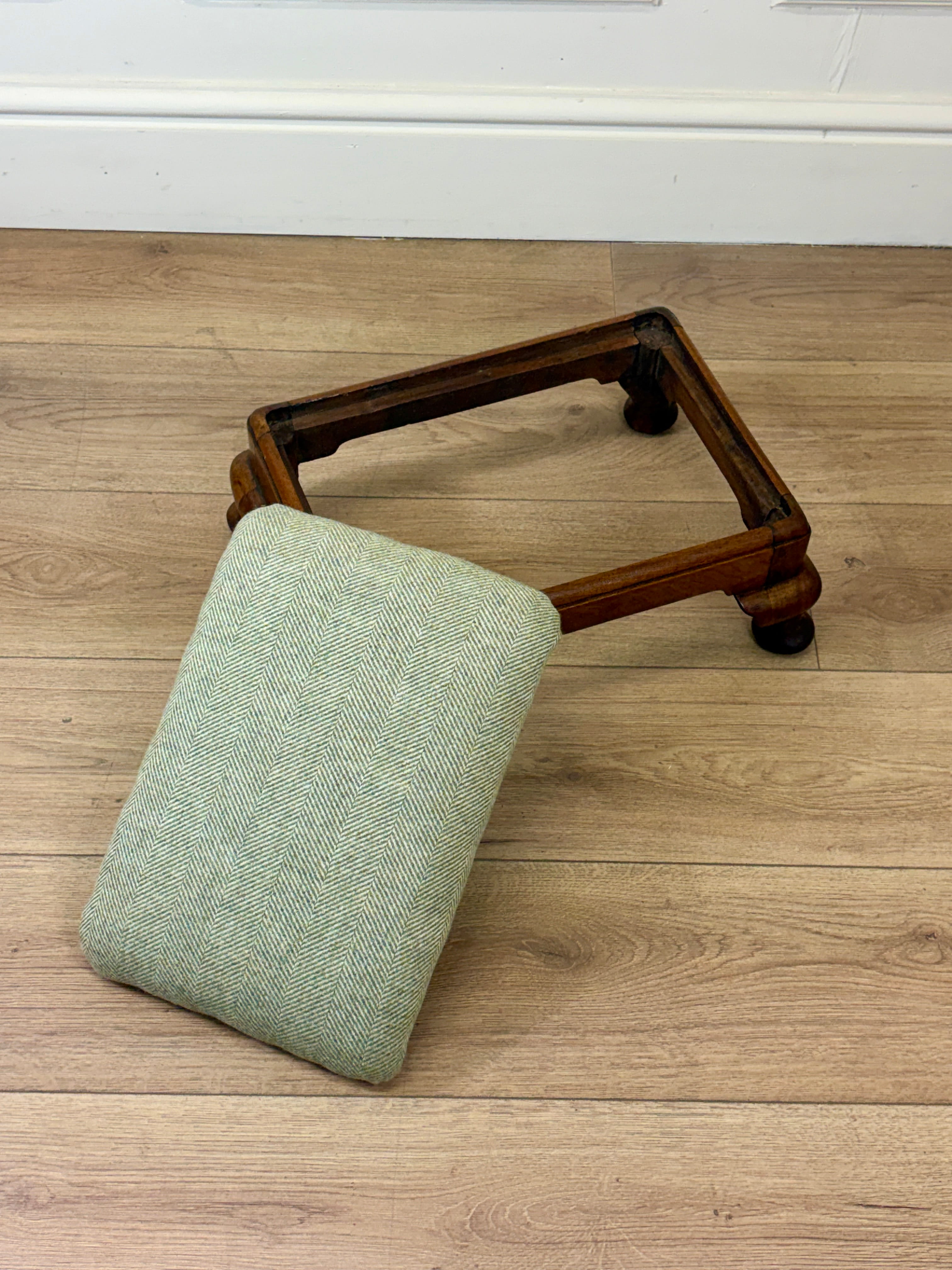 A small upholstered cushion in a light sage green fabric rests against a dark wood rectangular frame on a light oak floor.