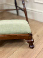 A close-up of a wooden stool or bench with a light sage green herringbone upholstered seat cushion, set on a light wood floor.