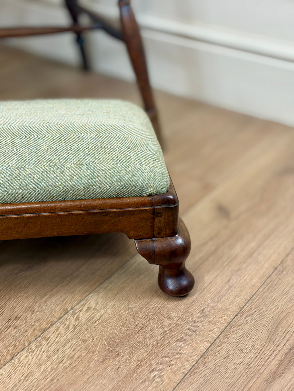 Close-up of a wooden stool or ottoman with a light sage green herringbone upholstered seat cushion, shown on a light wood floor in an indoor room setting.