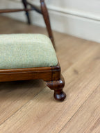 Close-up of a wooden stool or ottoman with a light sage green herringbone upholstered seat cushion, shown on a light wood floor in an indoor room setting.