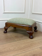 A small wooden footstool with a light sage green herringbone upholstered cushion and turned dark brown wooden legs, set on a light wood floor against a white paneled wall.