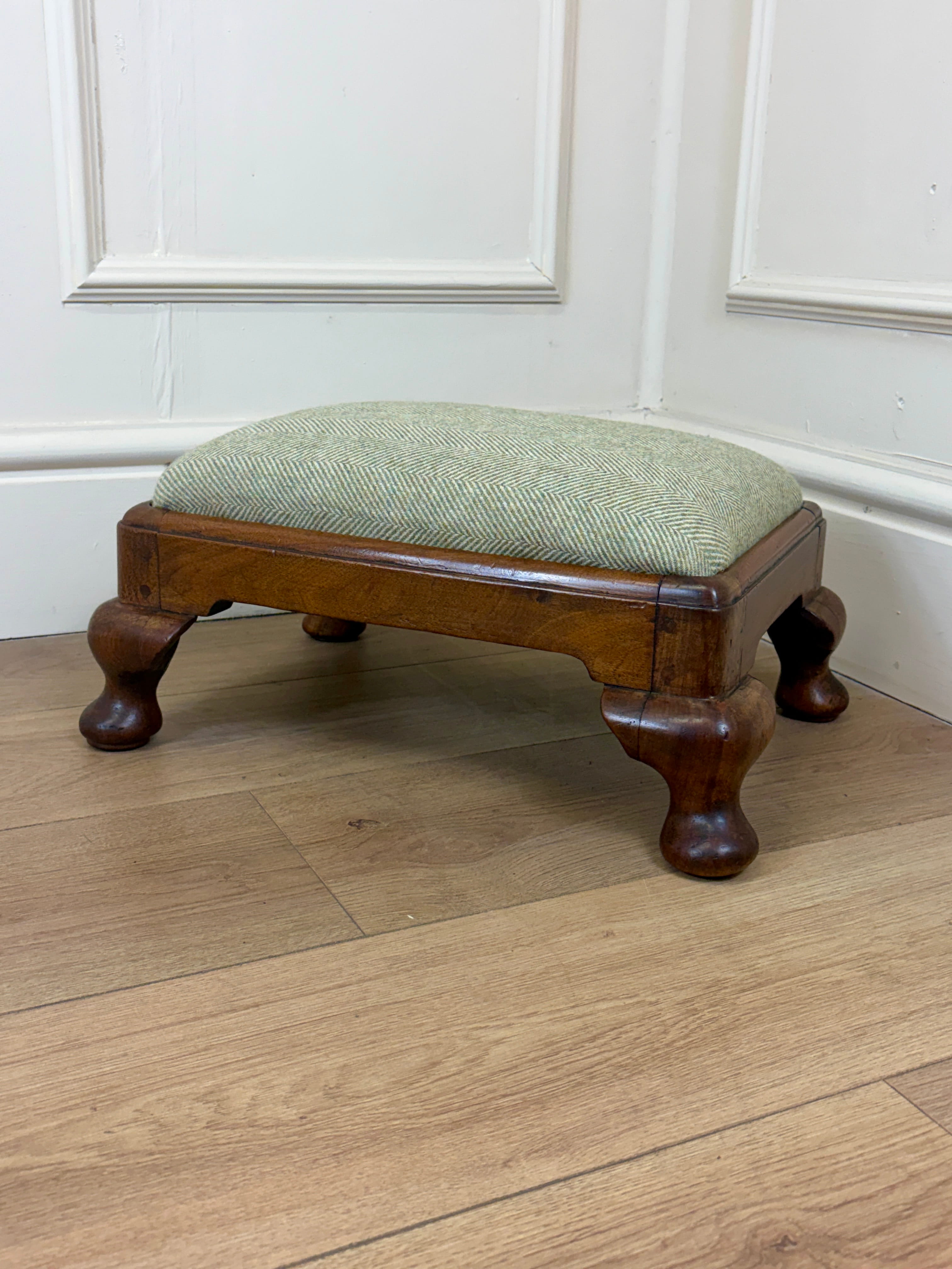 A small wooden footstool with a light sage green herringbone upholstered cushion and turned dark brown wooden legs, set on a light wood floor against a white paneled wall.