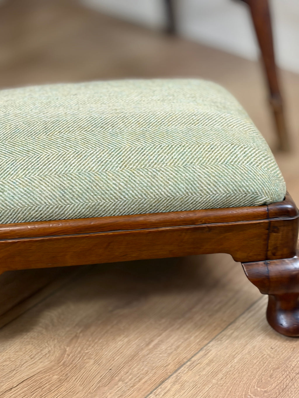 A close-up of a wooden stool or ottoman with a light sage green and beige herringbone upholstered cushion, resting on a light wood floor.