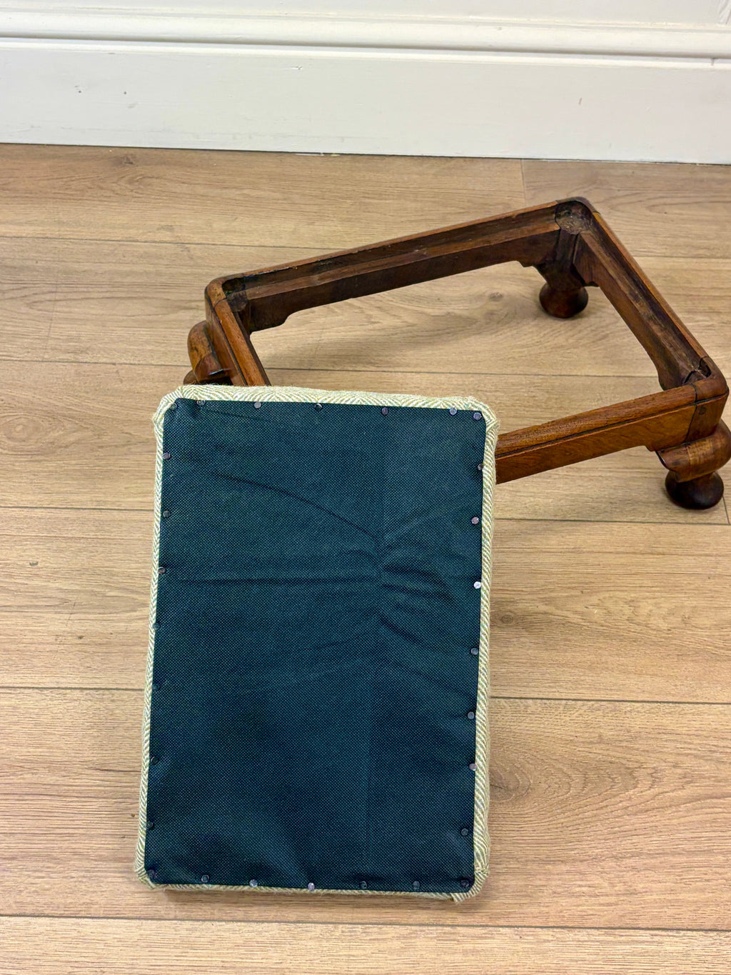 A dark teal fabric-covered rectangular cushion or pad with a light beige stitched border, placed in front of a wooden frame on a light wood floor.