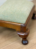 Close-up of a wooden stool or bench with a light green herringbone upholstered seat cushion and turned wood legs on a light wood floor.