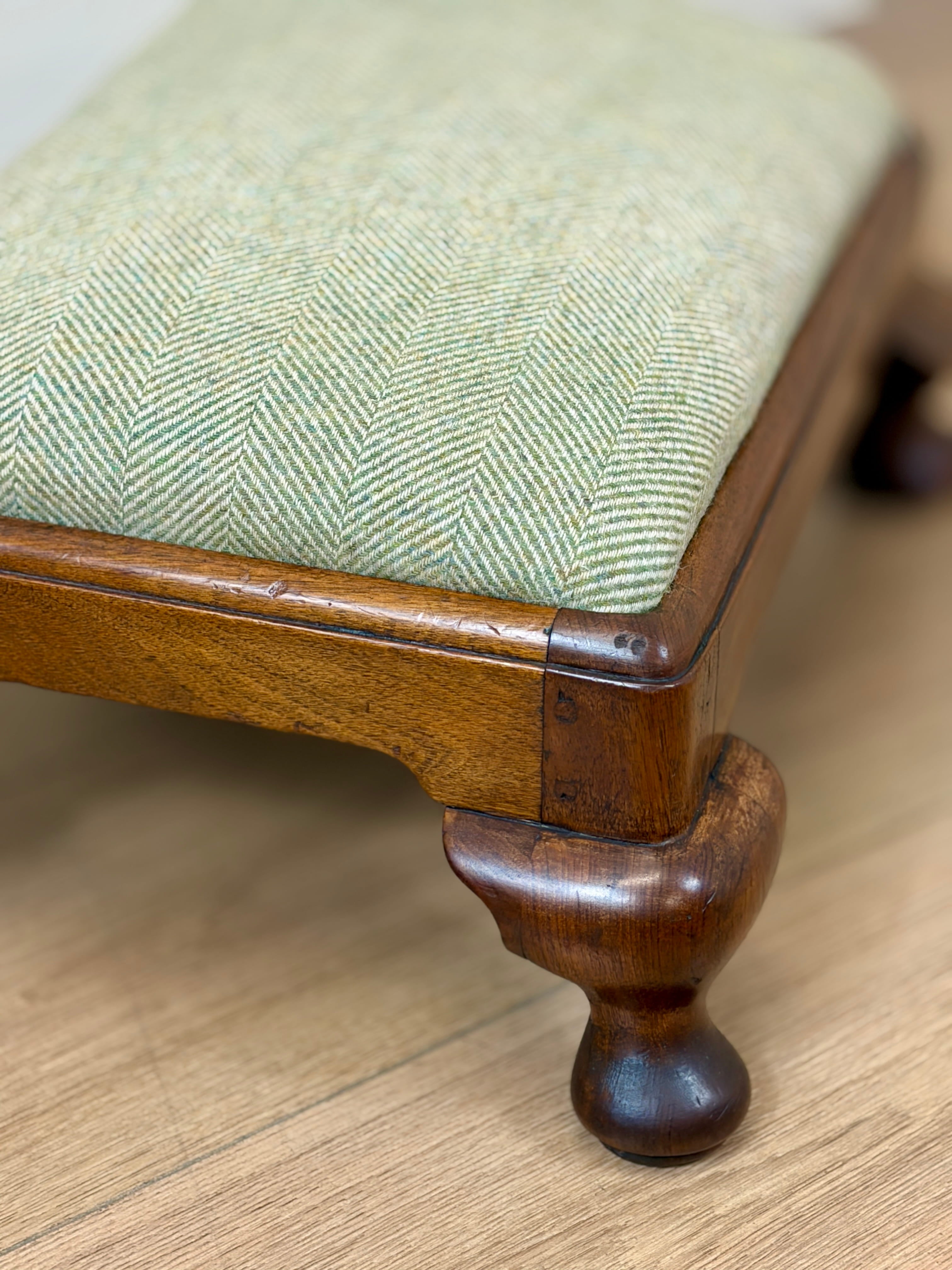 Close-up of a wooden stool or bench with a light green herringbone upholstered seat cushion and turned wood legs on a light wood floor.