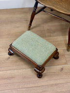 A small wooden stool with a light sage green herringbone upholstered cushion, shown on a light wood floor next to a larger wooden table.
