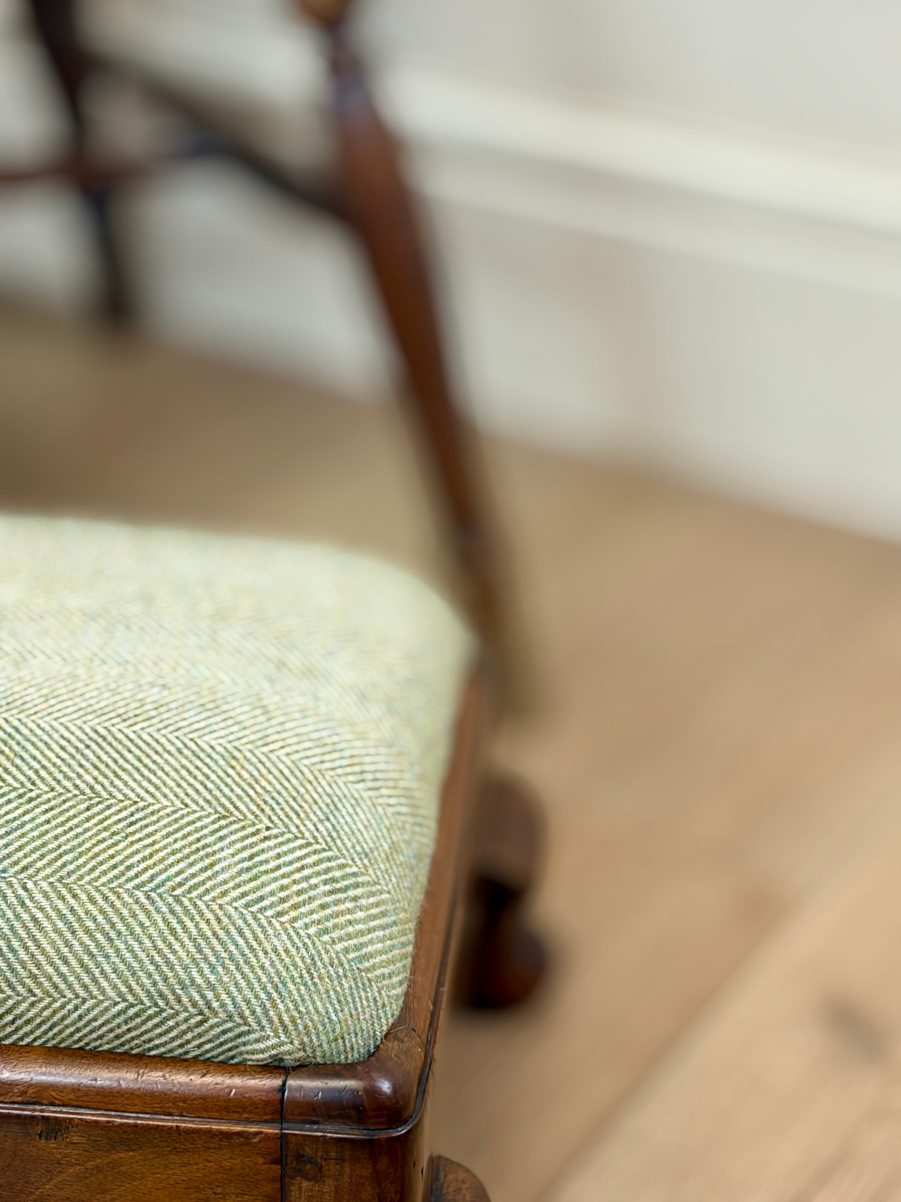 Close-up of a wooden stool or bench seat upholstered in a light sage green and cream herringbone fabric, with a warm wood frame and a softly blurred indoor background.