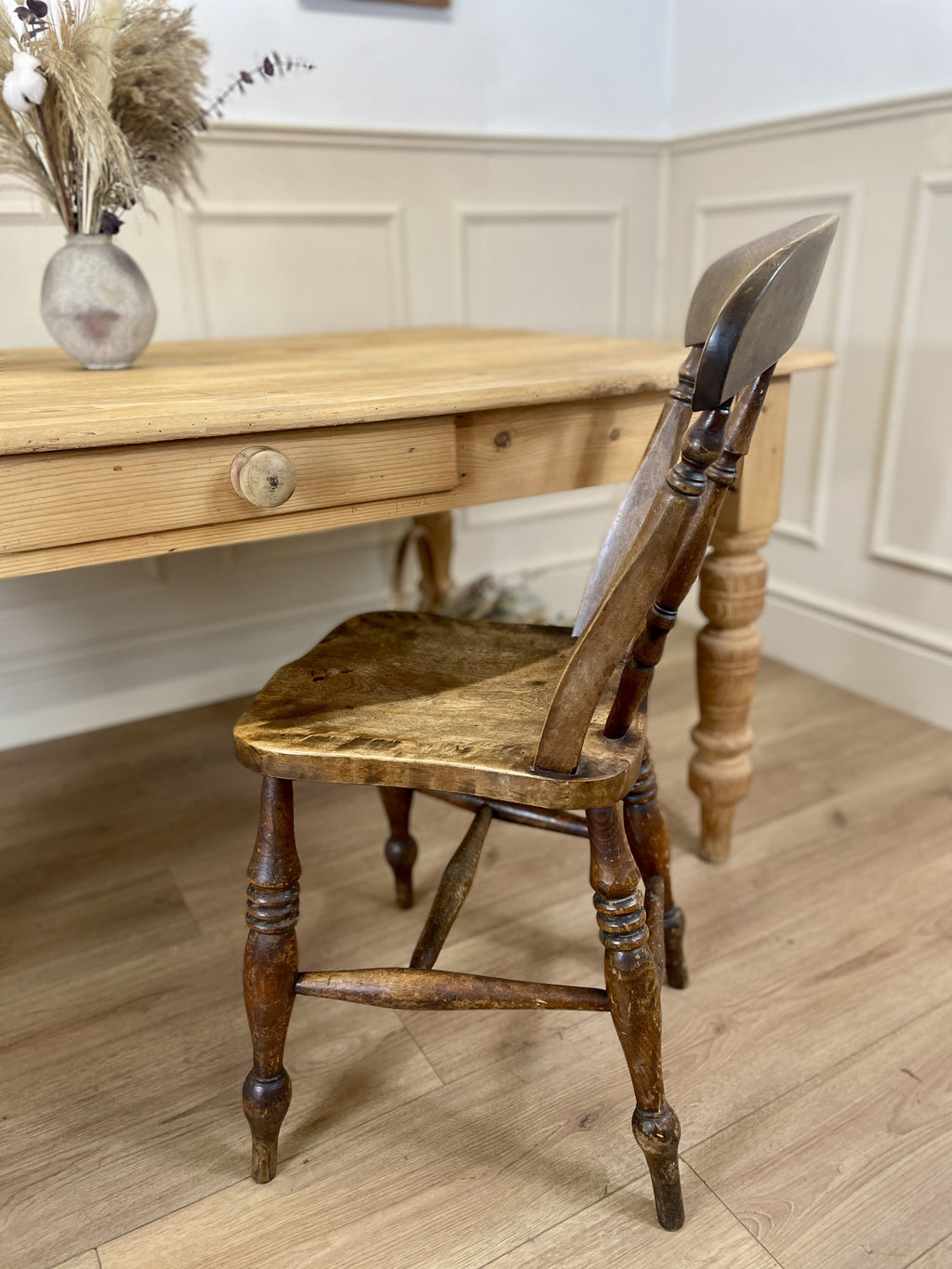 Wooden chair in front of a wooden table with a vase on a white paneled wall.