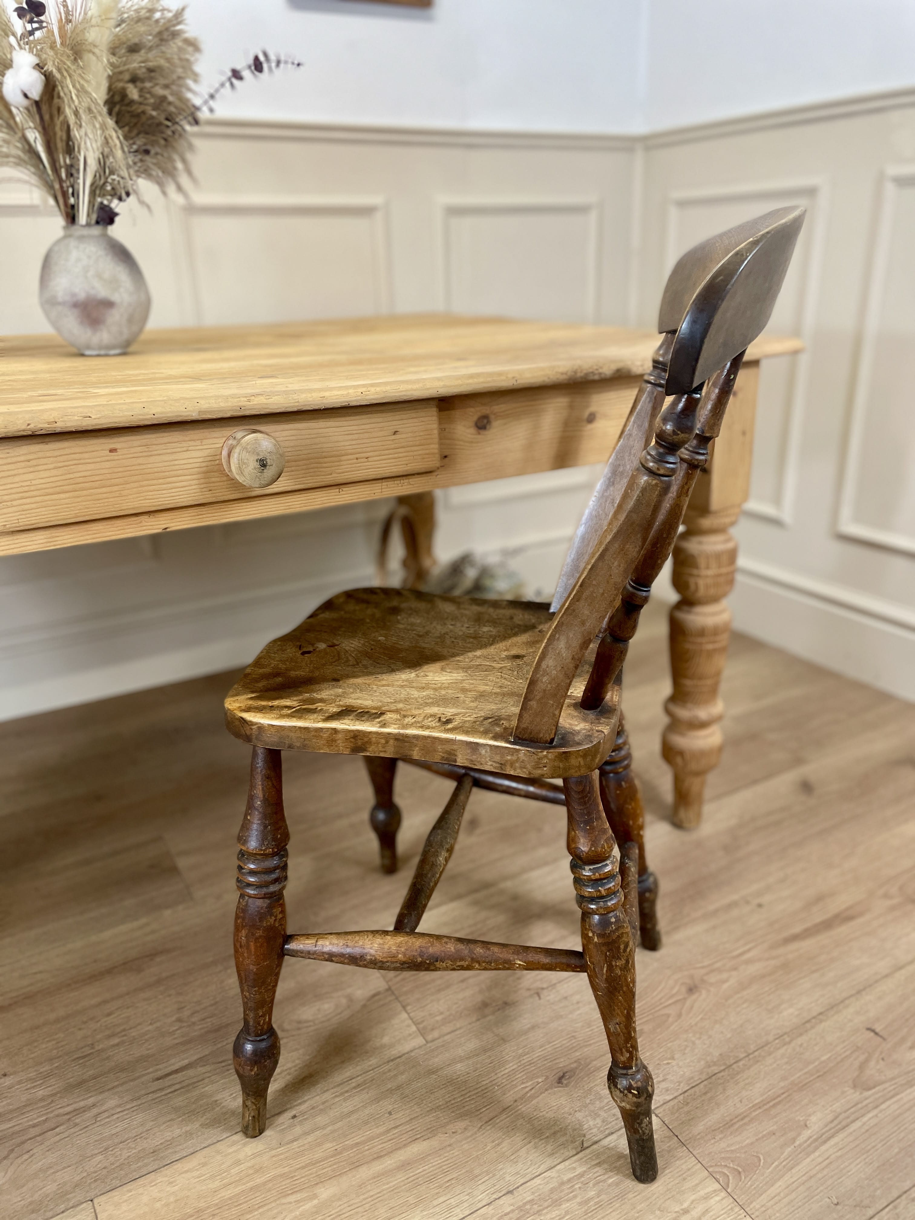 Wooden chair in front of a wooden table with a vase on a white paneled wall.
