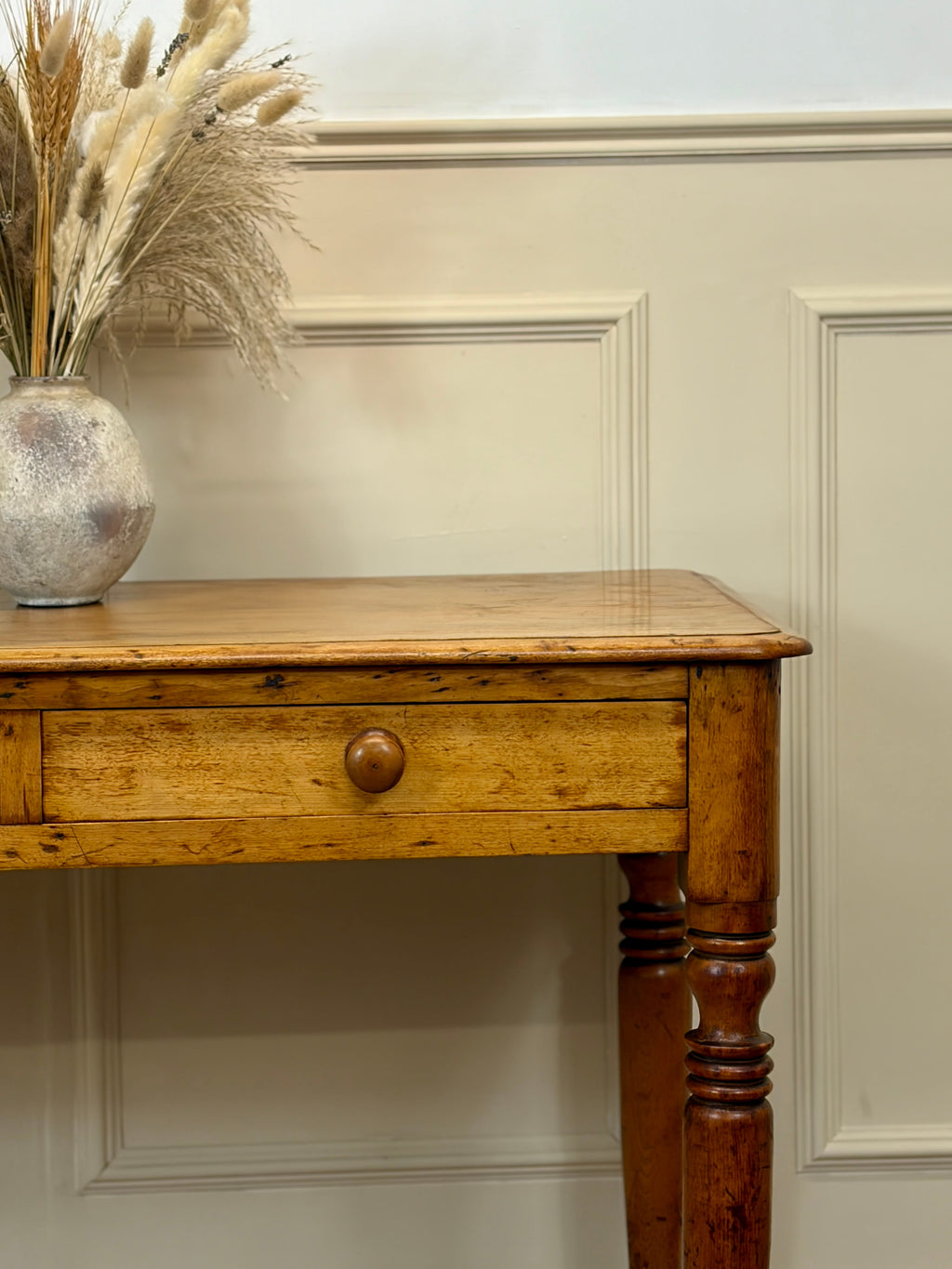 Wooden console table with a vase of dried plants against a paneled wall.