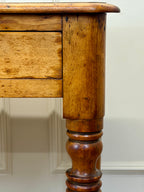 Close-up of a wooden desk with a drawer and turned leg against a neutral wall.