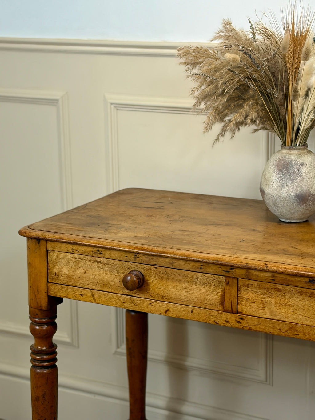 Wooden table with a vase of dried plants against a white paneled wall