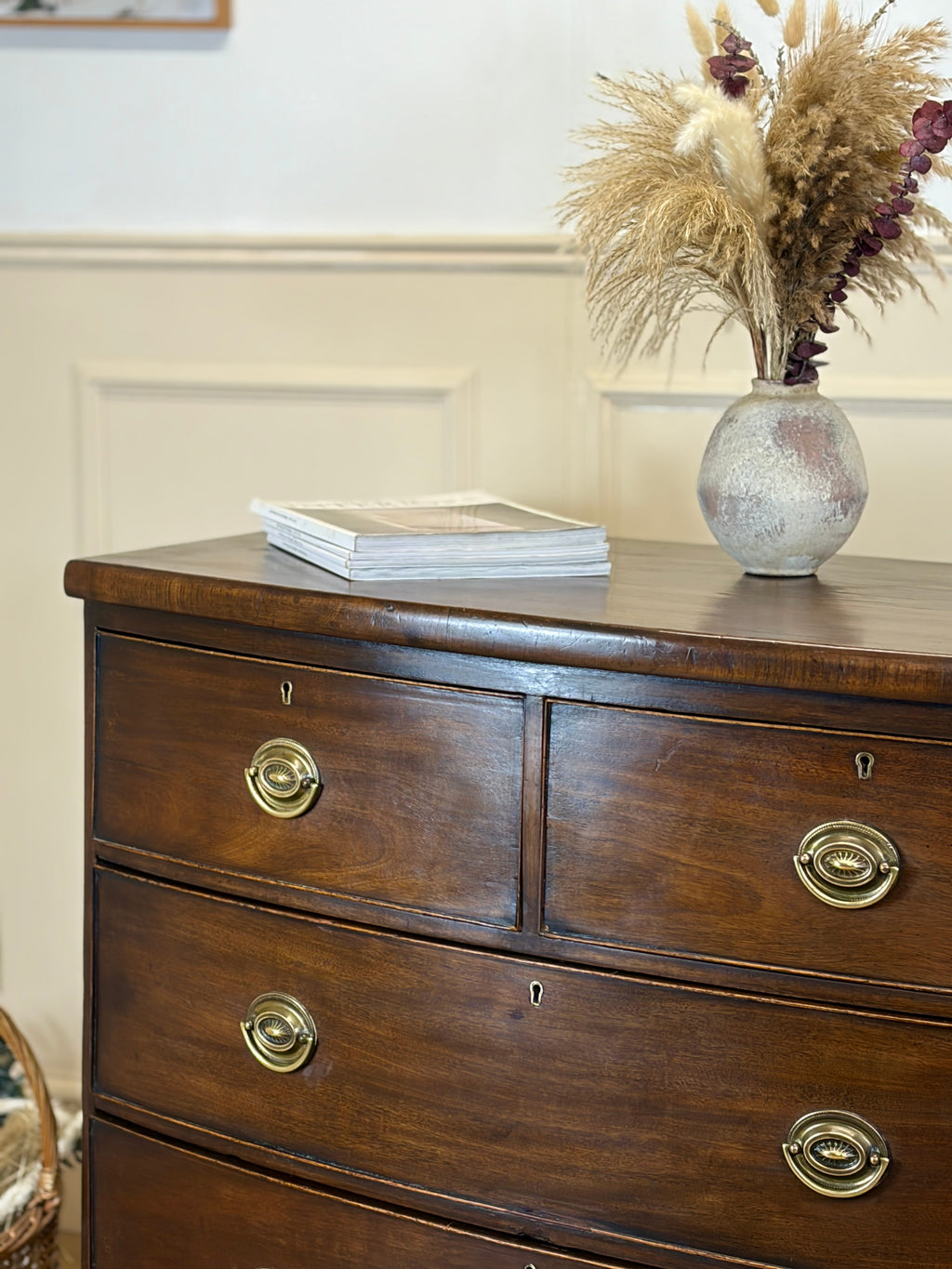 Wooden dresser with brass handles featuring a vase with dried plants and books on top.