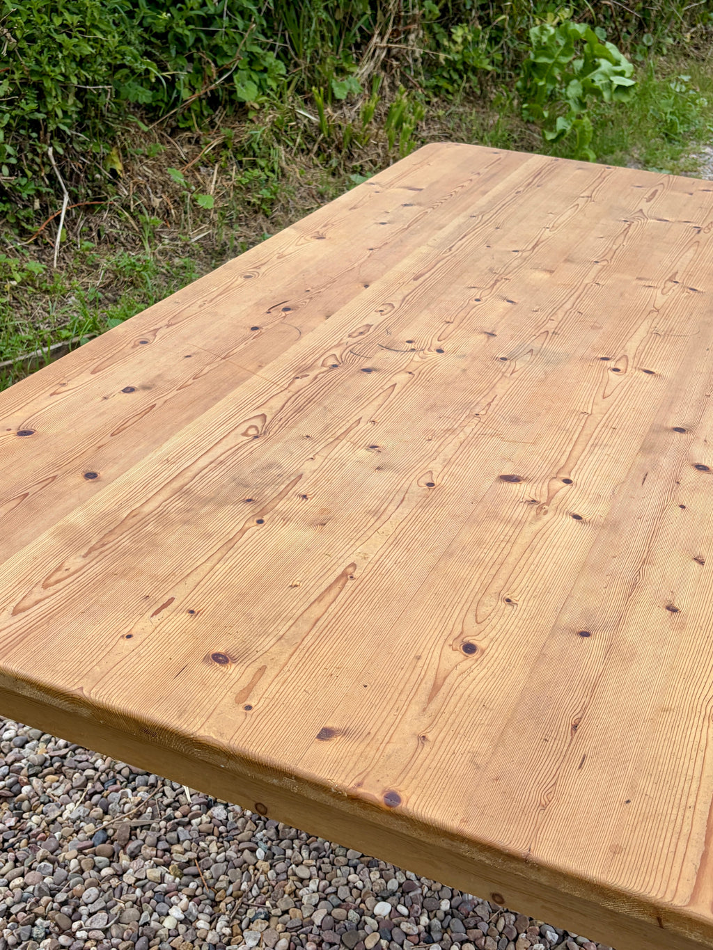 Wooden table top with natural grain and texture, outdoors on a gravel surface with greenery.