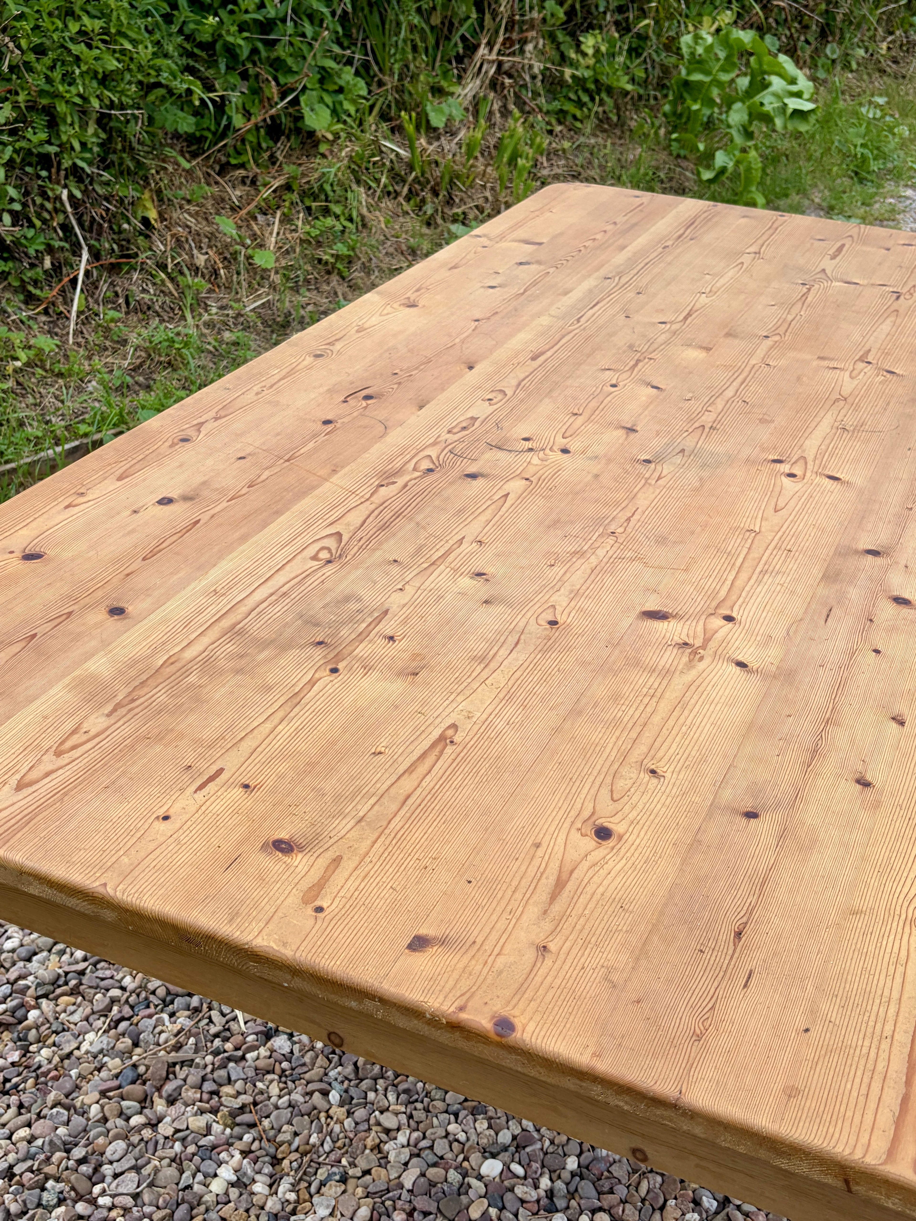 Wooden table top with natural grain and texture, outdoors on a gravel surface with greenery.