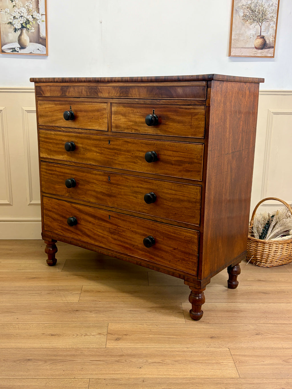 Wooden dresser with four drawers on a wooden floor, with a basket and framed pictures in the background.