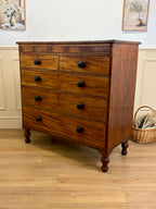 Wooden dresser with four drawers on a wooden floor, with a basket and framed pictures in the background.