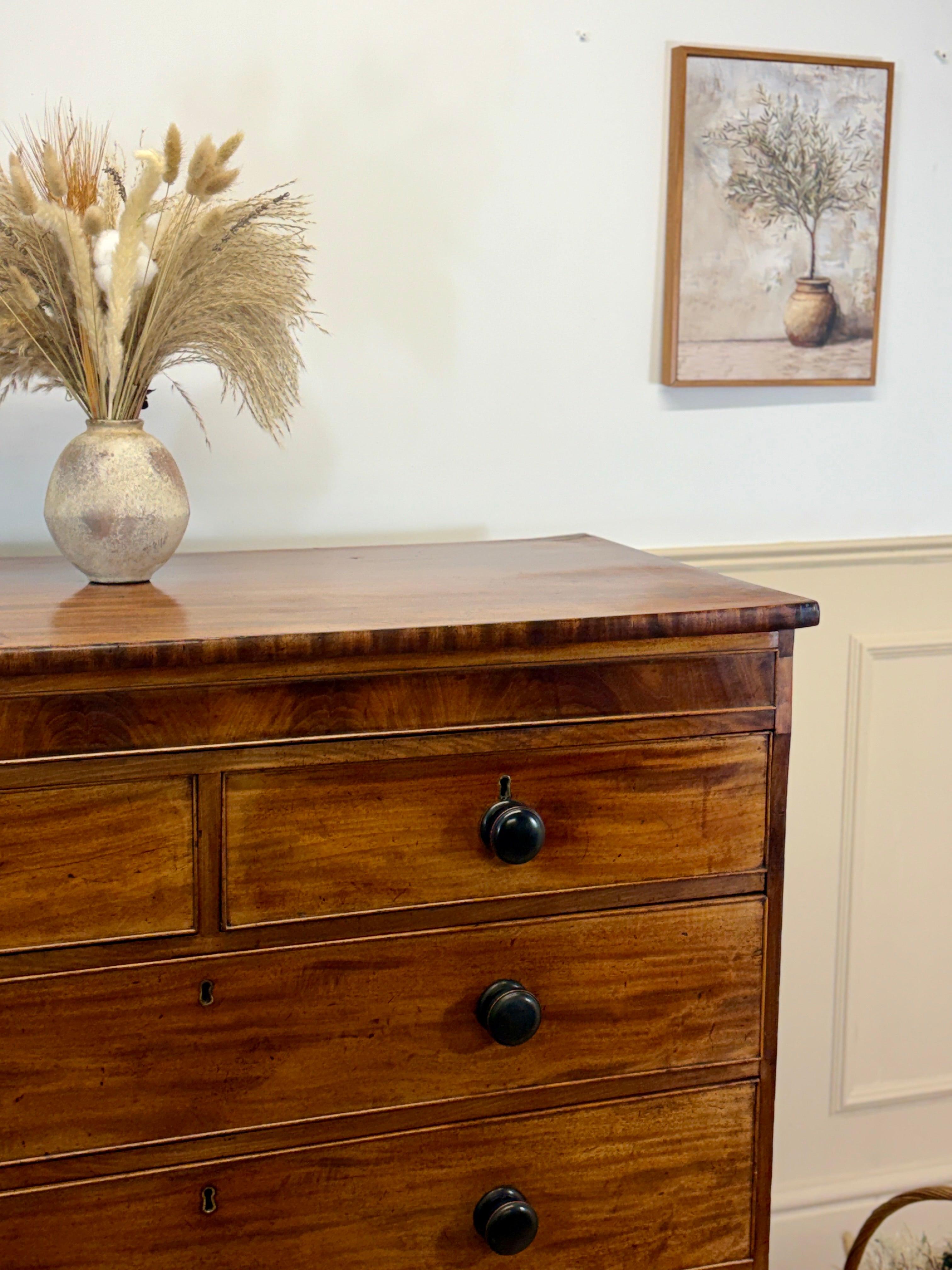 Wooden dresser with black knobs, vase of dried plants, and framed picture on a white wall.