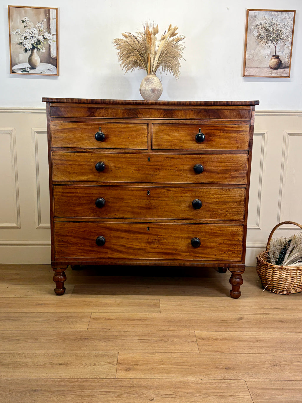 Wooden dresser with black knobs on a wooden floor, with two framed pictures on the wall.