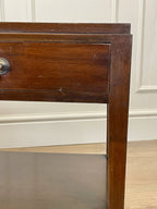 Wooden side table with a drawer against a white paneled wall.