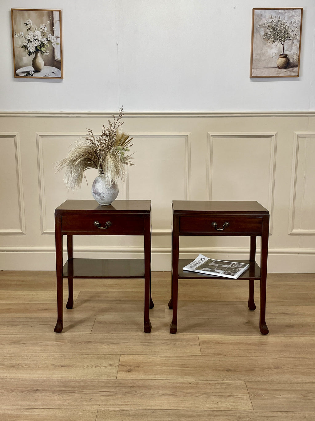 Two wooden side tables with a vase and newspaper against a wall with framed pictures.