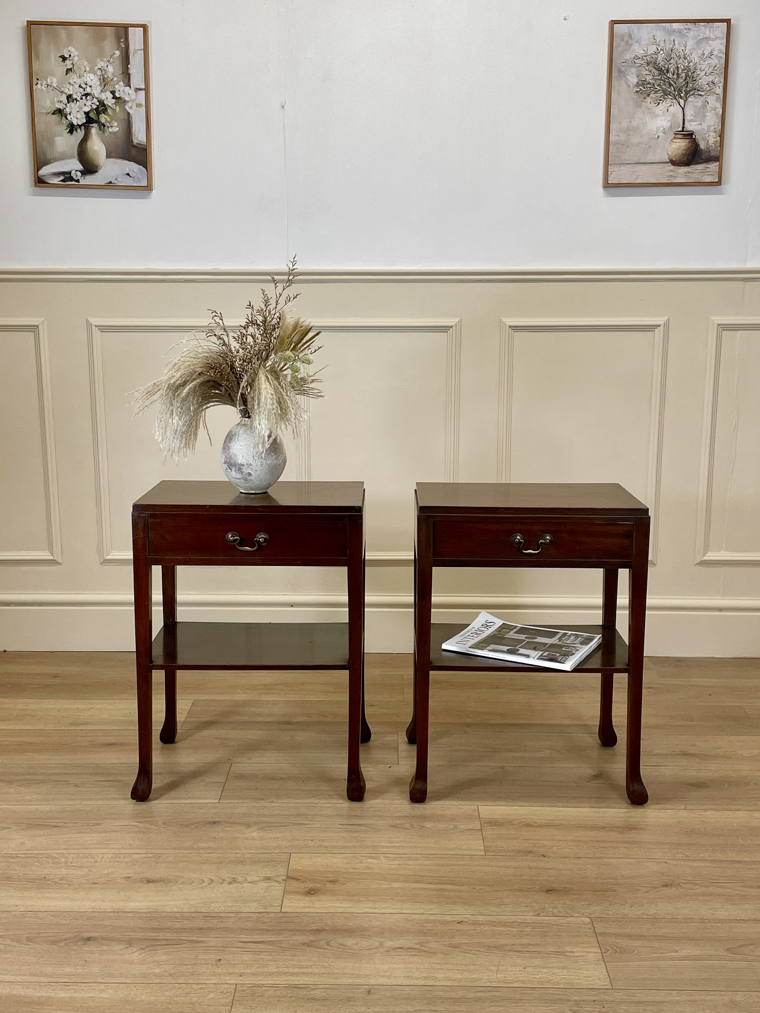 Two wooden side tables with a vase and newspaper against a wall with framed pictures.