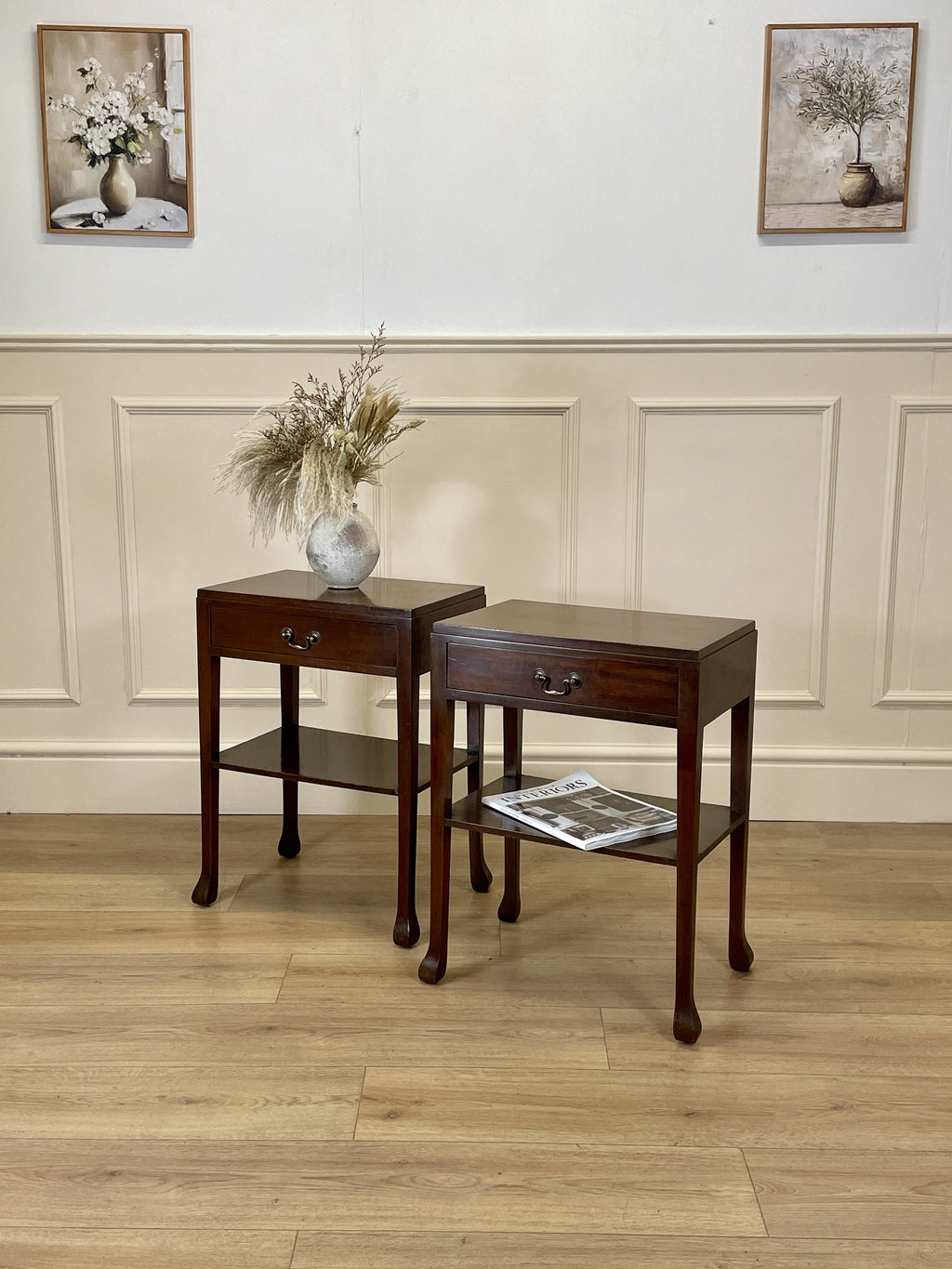 Two wooden side tables with a vase and magazine against a wall with framed pictures.