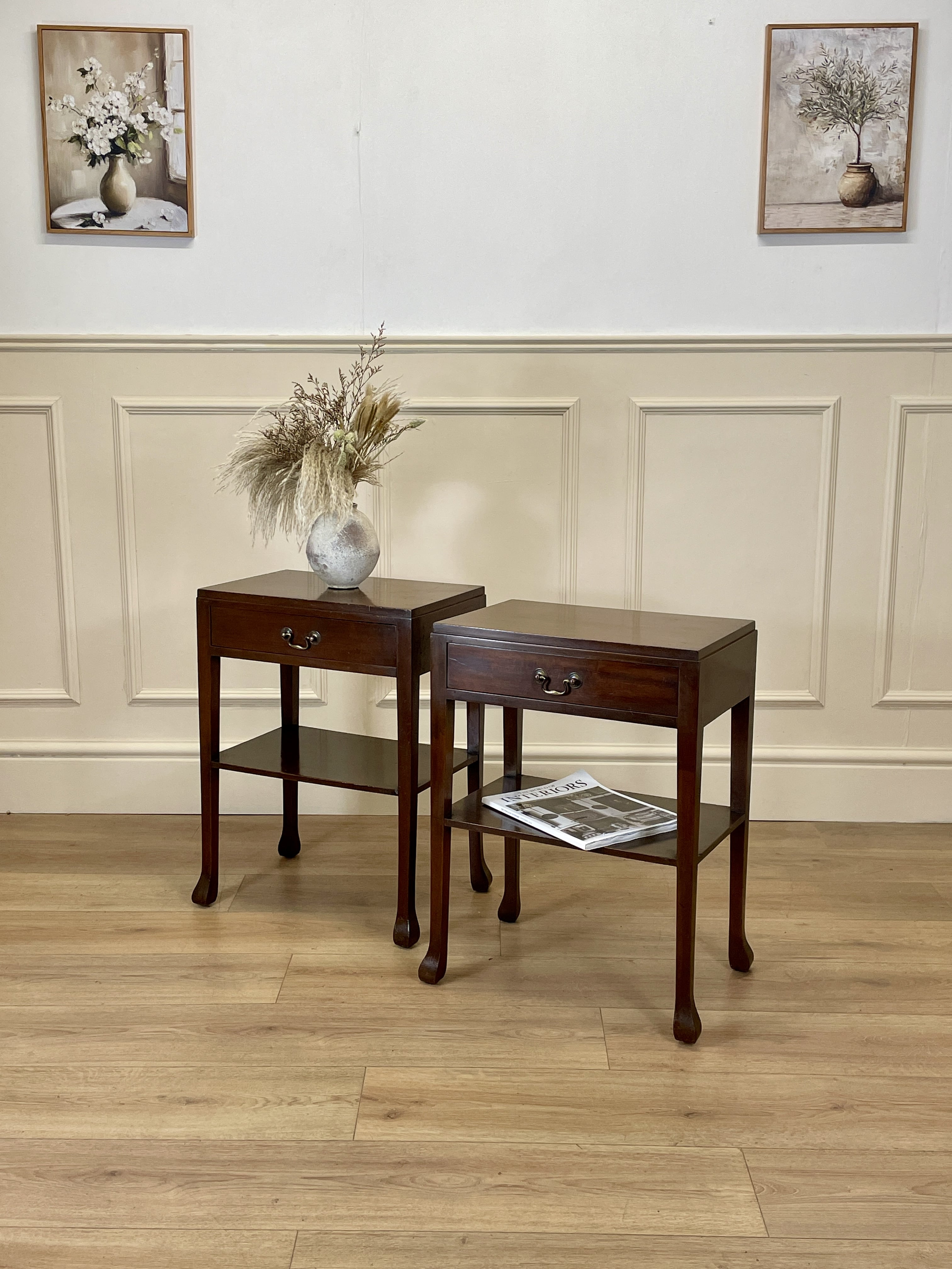 Two wooden side tables with a vase and magazine against a wall with framed pictures.