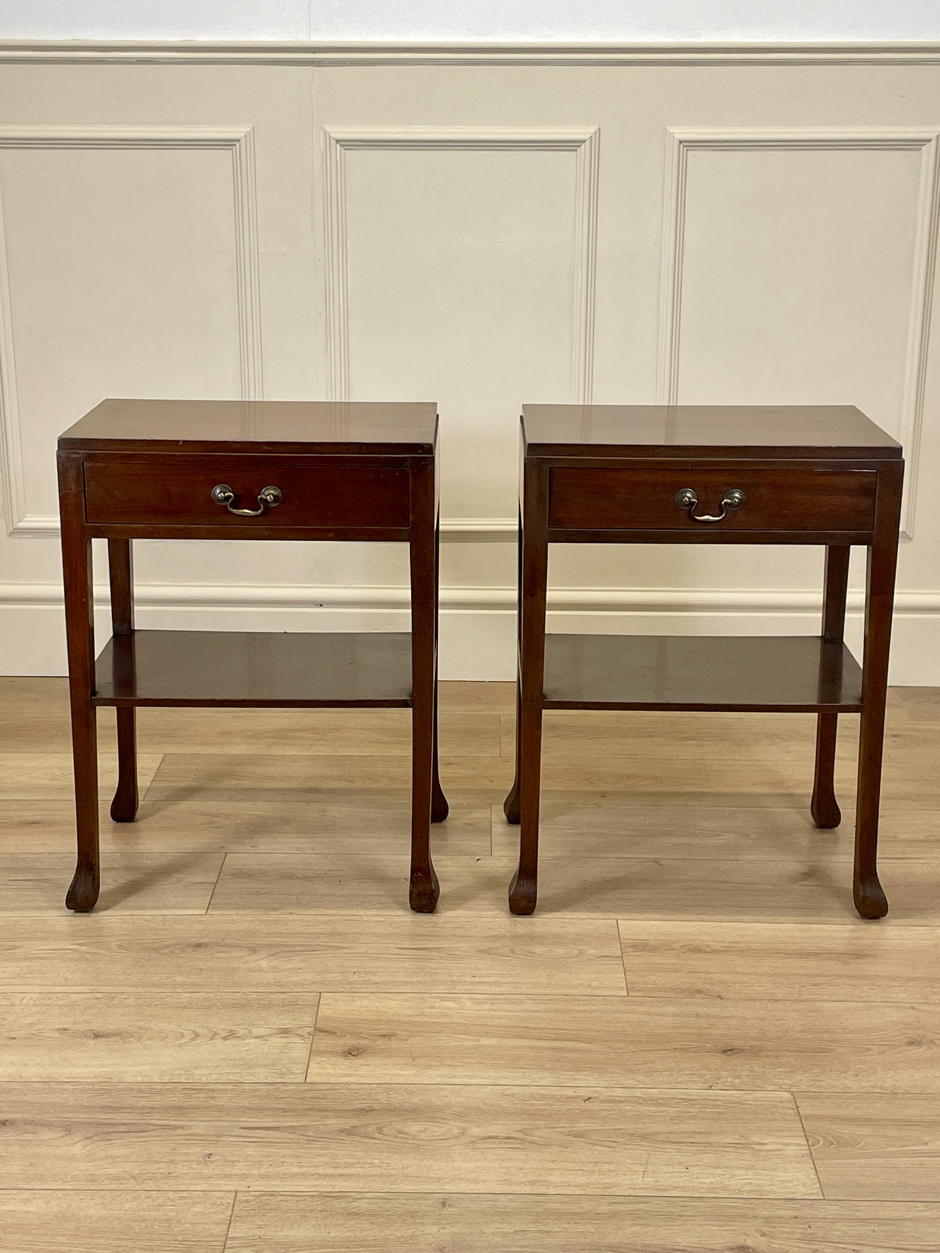 Two wooden side tables with drawers on a wooden floor against a paneled wall.
