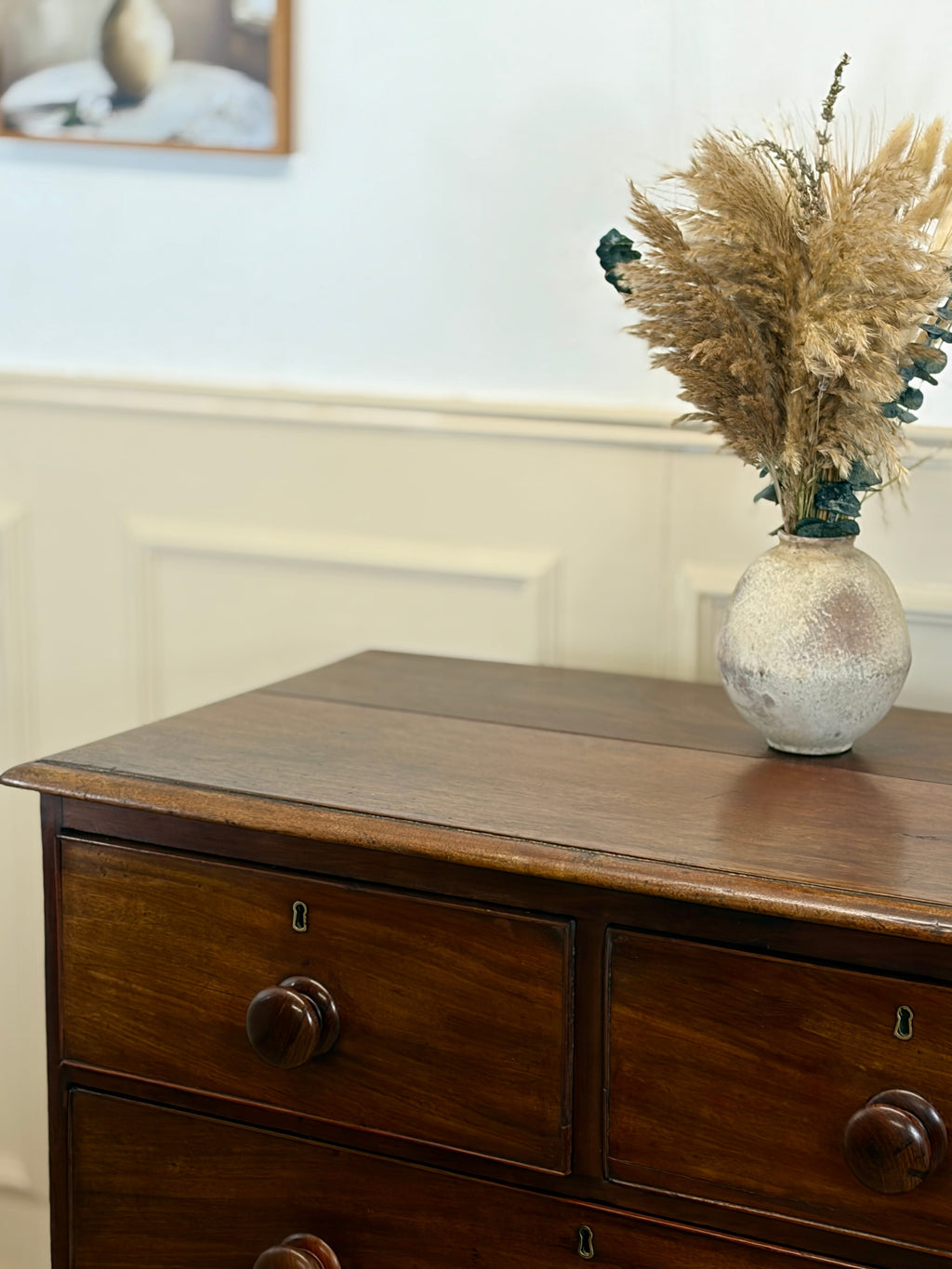 Wooden dresser with a vase of dried pampas grass on top, against a light wall.