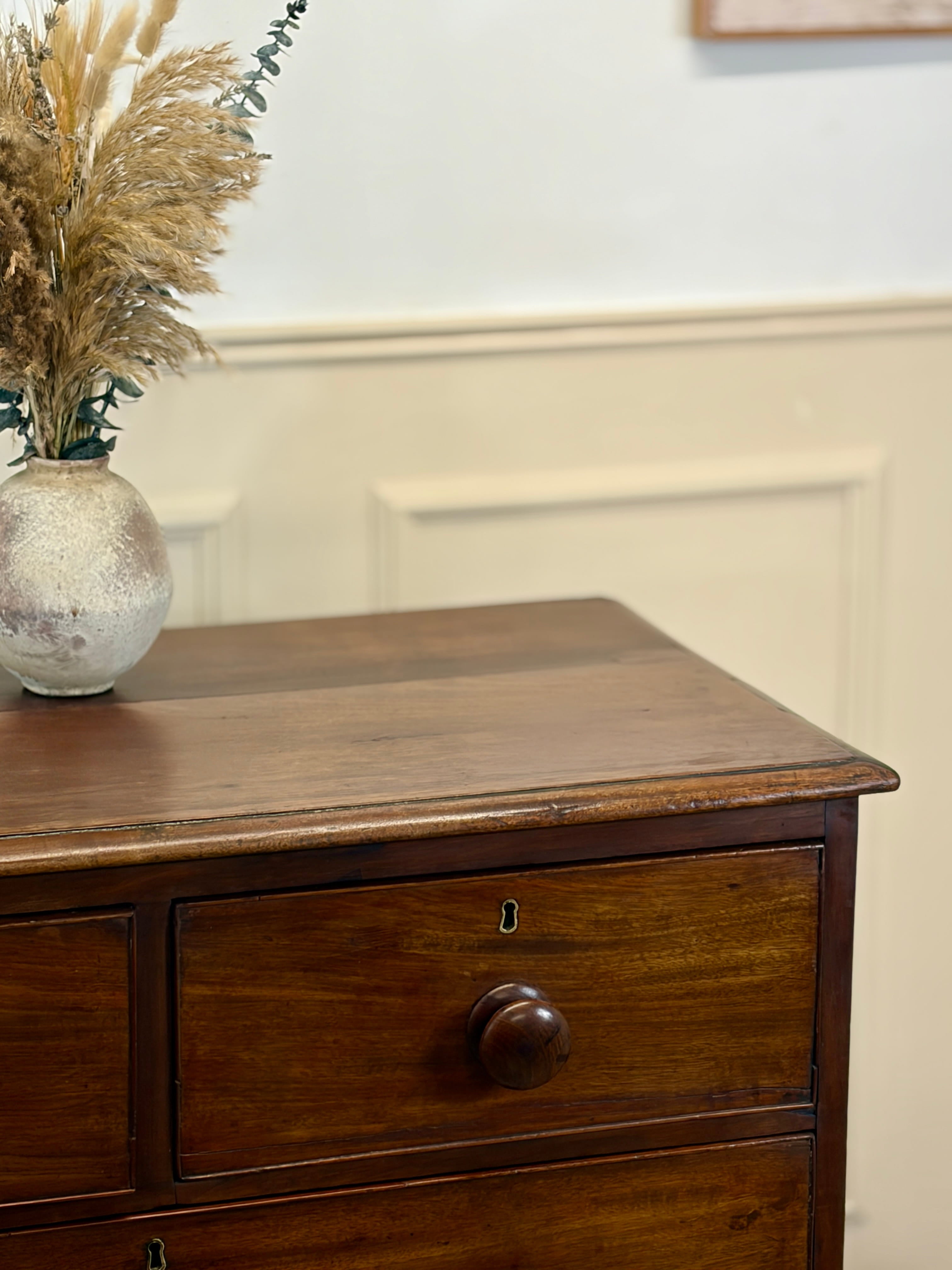 Wooden dresser with a vase of dried plants on top against a beige wall.
