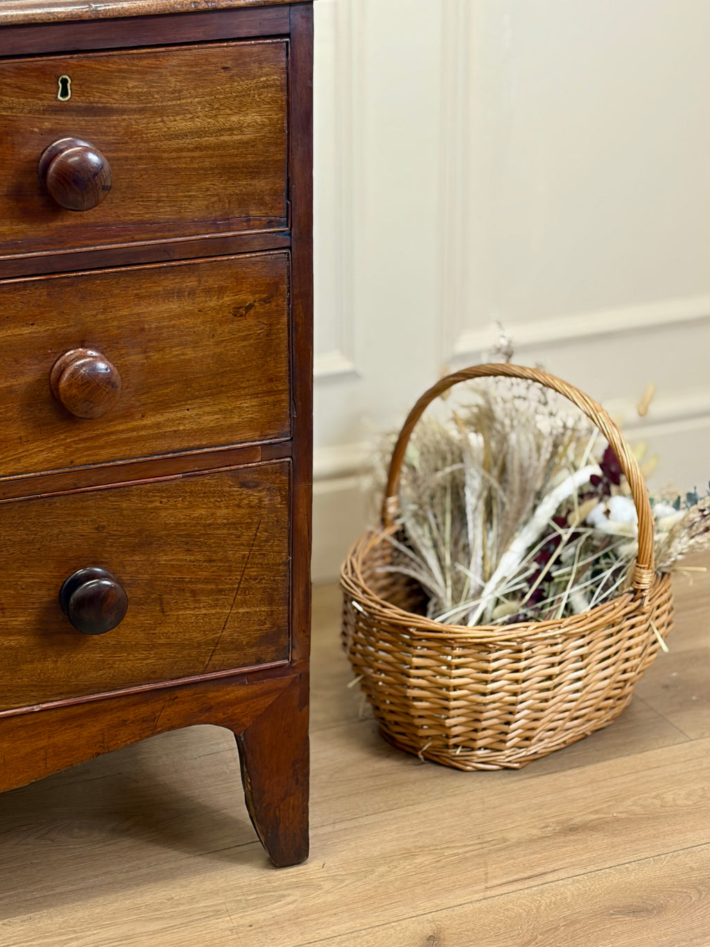 Wooden dresser with wicker basket containing dried plants on a wooden floor.