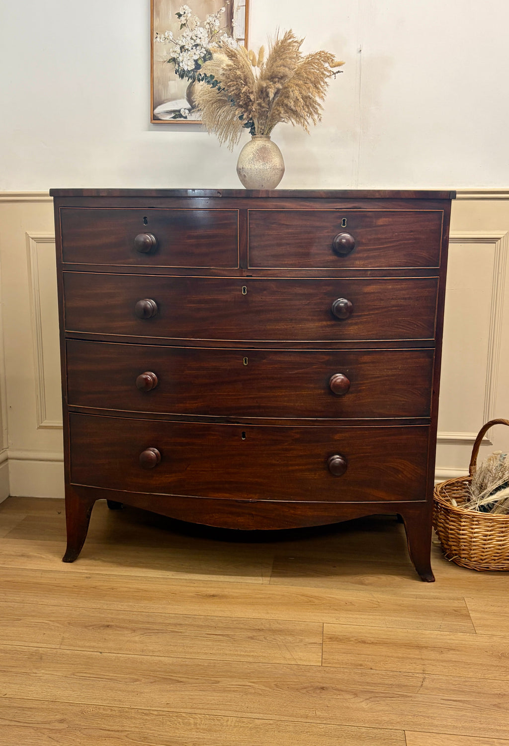 Wooden dresser with four drawers on a wooden floor, with a vase of flowers and a basket in the background.