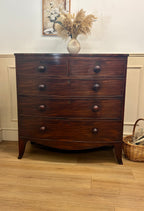 Wooden dresser with four drawers on a wooden floor, with a vase of flowers and a basket in the background.