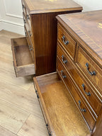 Vintage pair of mahogany bedside chest of drawers with five drawers each, featuring decorative inlaid tops and brass handles, displayed against a cream wall on a wooden floor.