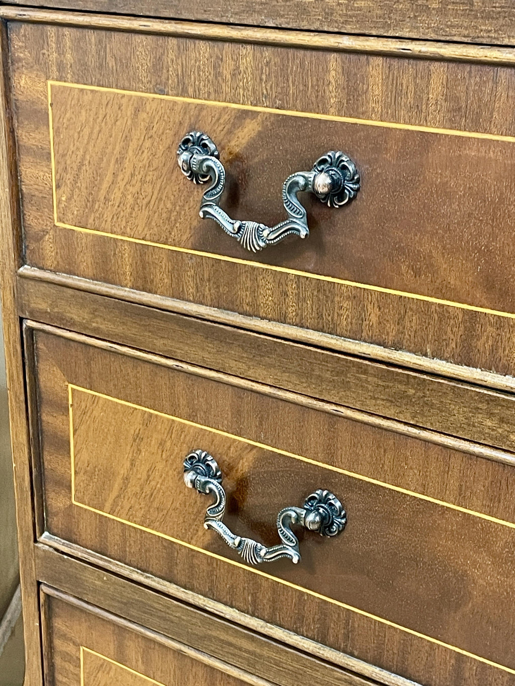 Vintage pair of mahogany bedside chest of drawers with five drawers each, featuring decorative inlaid tops and brass handles, displayed against a cream wall on a wooden floor.